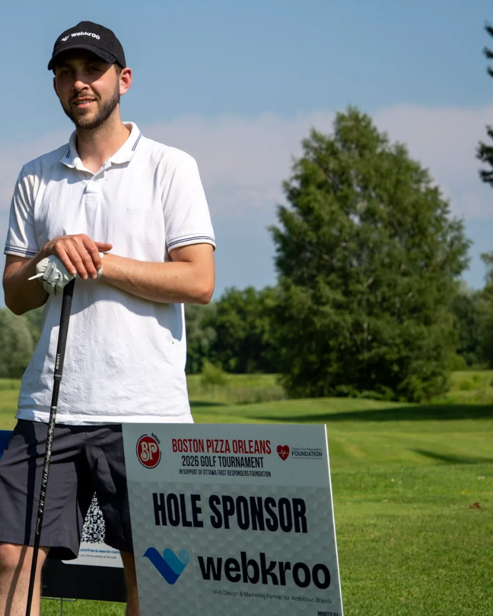 A golfer beside WebKroo's hole sponsor sign at the tournament tee.